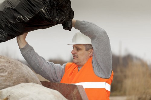 A male waste collection worker wearing a white safety helmet and an orange high-visibility vest is outdoors, lifting a large black rubbish bag above his head. He appears focused on the task, with a neutral expression. The background shows a blurred, overcast sky and a rural or suburban landscape with dry grass and sparse trees. In the foreground, he is standing beside a pile of mixed waste, including large, durable bags and various discarded materials. The scene is typical of a rubbish removal operation in the Bromley area, emphasizing the manual handling of bulky waste items. The worker's clothing and safety gear are appropriate for waste collection activities, and the environment looks like an open outdoor space suited for rubbish clearance services provided by Garden Clearance Bromley.