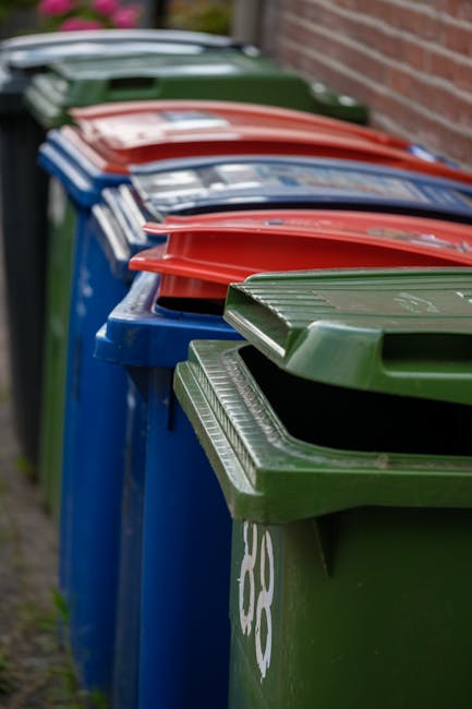 A row of large wheelie bins lined up along a paved pathway beside a brick wall on the right. The bins are made of plastic with textured lids, painted in various colors: the nearest bin in green, follo